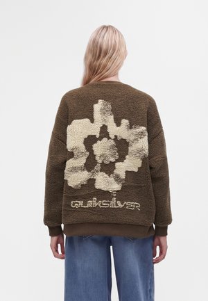Personne aux longs cheveux blonds, vue de dos, portant une veste polaire marron avec un grand motif floral crème et le texte "quiksilver" dans le dos.