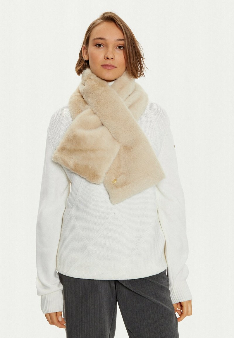 Sciarpa in pelliccia sintetica beige con una texture morbida, decorata con un accento dorato. Indossata in modo rilassato attorno al collo sopra un maglione bianco lavorato a maglia.