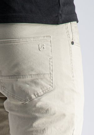 Primer plano del bolsillo de unos pantalones beige claro con un logotipo sutil bordado, combinados con una camisa negra metida dentro a la altura de la cintura.