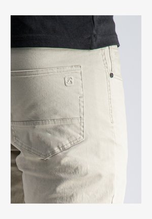 Primer plano del bolsillo de unos pantalones beige claro con un logotipo sutil bordado, combinados con una camisa negra metida dentro a la altura de la cintura.