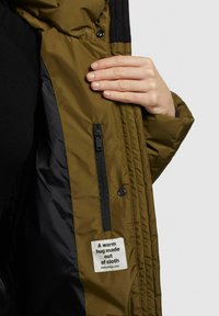 Olijfgroene pufferjack interieur met zwarte voering, ritszak en label met de tekst "Een warme omhelzing gemaakt van stof."