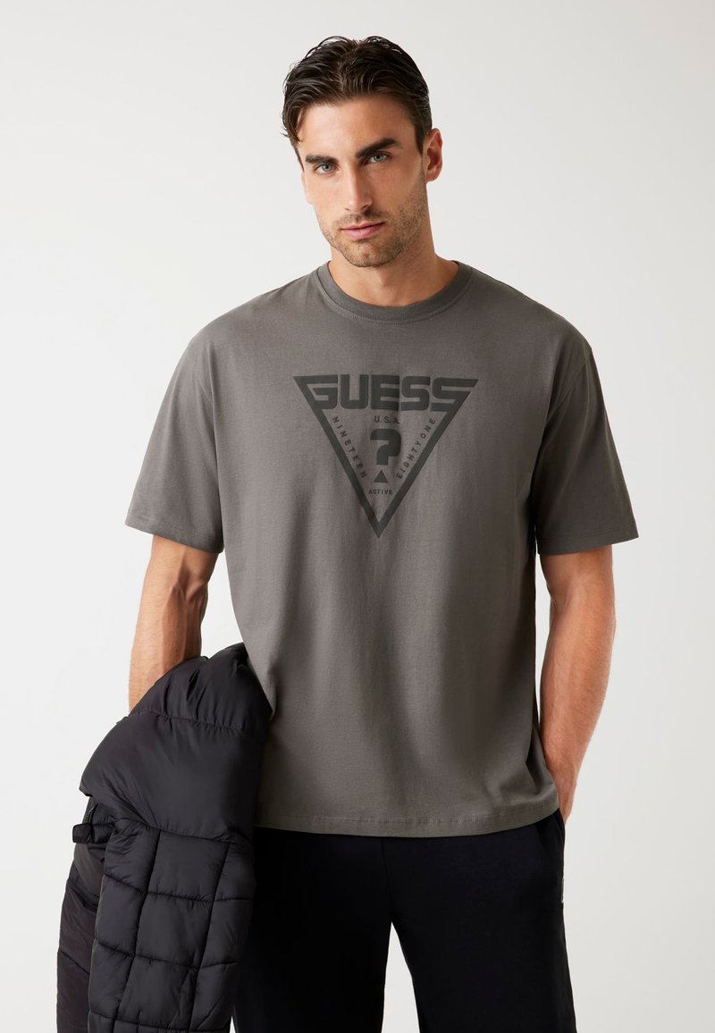 Hombre vistiendo una camiseta gris de Guess con el logo, sosteniendo una chaqueta de plumas negra con una mano, de pie contra un fondo blanco plano.