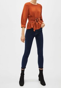 Kvinde iført en struktureret, brændt orange langærmet bluse med bælte, mørkeblå skinny jeans og sorte hælede ankelstøvler, der står foran en hvid baggrund.