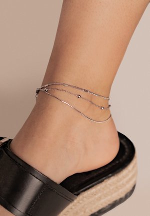 Silver anklet with multiple layered chains, featuring varied textures and small beads. Worn with a black slide sandal on a bare foot.