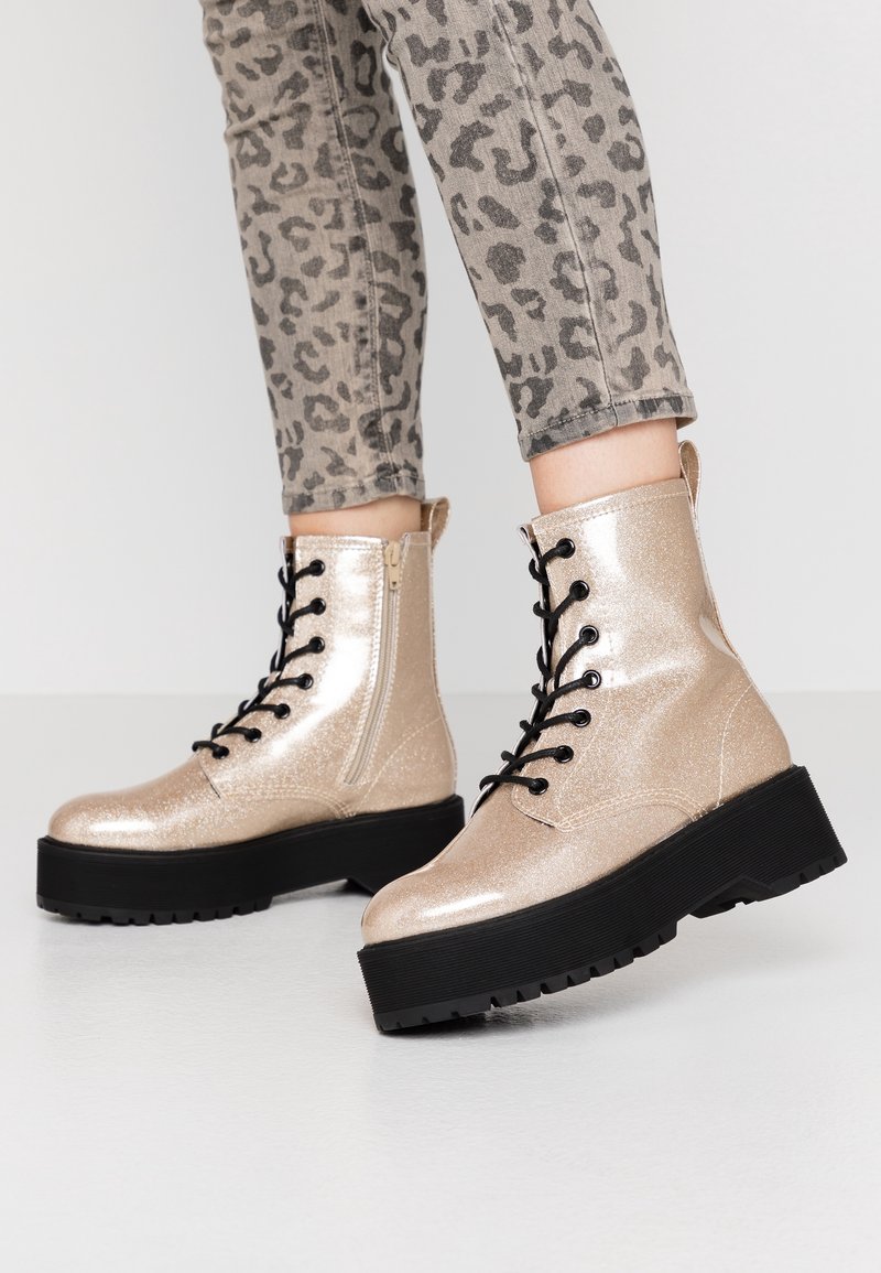 Metalen gouden combatlaarzen met zwarte veters, een zijrits en dikke zwarte platformzolen. Getextureerd oppervlak met een glinsterende finish.