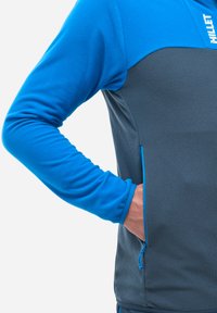 Veste en polaire présentant une partie supérieure bleu vif et une partie inférieure gris foncé. Tissu texturé avec une poche latérale zippée.