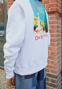 Bílá mikina s barevným grafickým vzorem, který zobrazuje věž a krajinu, s textem "Na dovolené" pod tímto designem. Hladké manžety a lem.