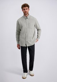 Camicia grigia chiaro con bottoni e taschino sul petto, abbinata a pantaloni scuri con texture e sneaker bianche con suola marrone. Sfondo semplice.