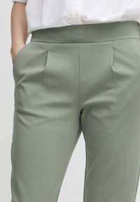 Jasnozielone spodnie o gładkiej fakturze, z plisami z przodu i bocznymi kieszeniami, zestawione z białą koszulą z guzikami.