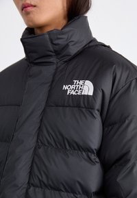 Giacca a vento nera con colletto alto, caratterizzata da una texture trapuntata e un importante logo bianco "The North Face" sul petto.