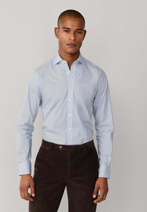 Camisa de botones celeste con un patrón sutil, de manga larga y con cuello, combinada con pantalones de pana marrón oscuro.