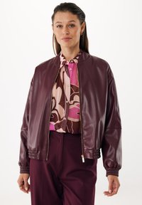 Giubbotto bomber in similpelle bordeaux con polsini a coste e chiusura con zip. Indossato sopra una blusa a stampa floreale rosa e marrone, creando un look a strati.