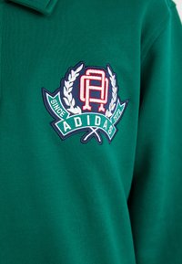 Grønn bomullspolo med et brodert logo med "ADIDAS", et dekorativt skjold og laurbærdekorasjoner. Glatt tekstur, kort krage.