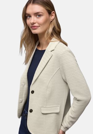 Blazer beige clair avec un motif texturé côtelé, fermeture à un bouton et poches latérales. Porté sur un haut bleu marine.