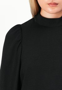 Kvinne ikledd en svart høyhalset topp med puffede ermer, som viser leppene og deler av det blonde håret mot en hvit bakgrunn.