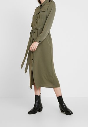 Mujer vistiendo un vestido midi de manga larga color verde oliva con cinturón y botones, con bolsillos en el pecho y botas negras hasta el tobillo, sobre un fondo liso.