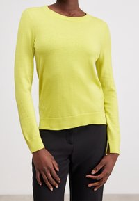 Bright yellow long-sleeve knit sweater with a rounded neckline, side slits, and ribbed cuffs, paired with black trousers.