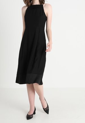 Kobieta w czarnej, bezrękawnikowej sukience midi z kwadratowym dekoltem i czarnych szpilkach z ozdobnymi wycięciami, stojąca na jednolitym tle.