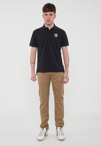 Maglietta polo blu navy con un logo circolare, pantaloni khaki e sneakers bianche con dettagli verdi. Il tessuto appare liscio; design aderente.