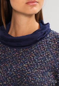 Marinblå texturerad topp med flerfärgade prickar; har en kontrasterande mörkblå cowl-hals. Tyget verkar mjukt och stickat.