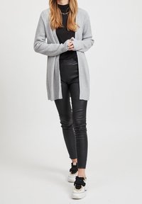 Cardigan tricoté gris clair avec un devant ouvert, texture côtelée, porté sur un top noir et un legging en cuir noir, associé à des baskets blanches.