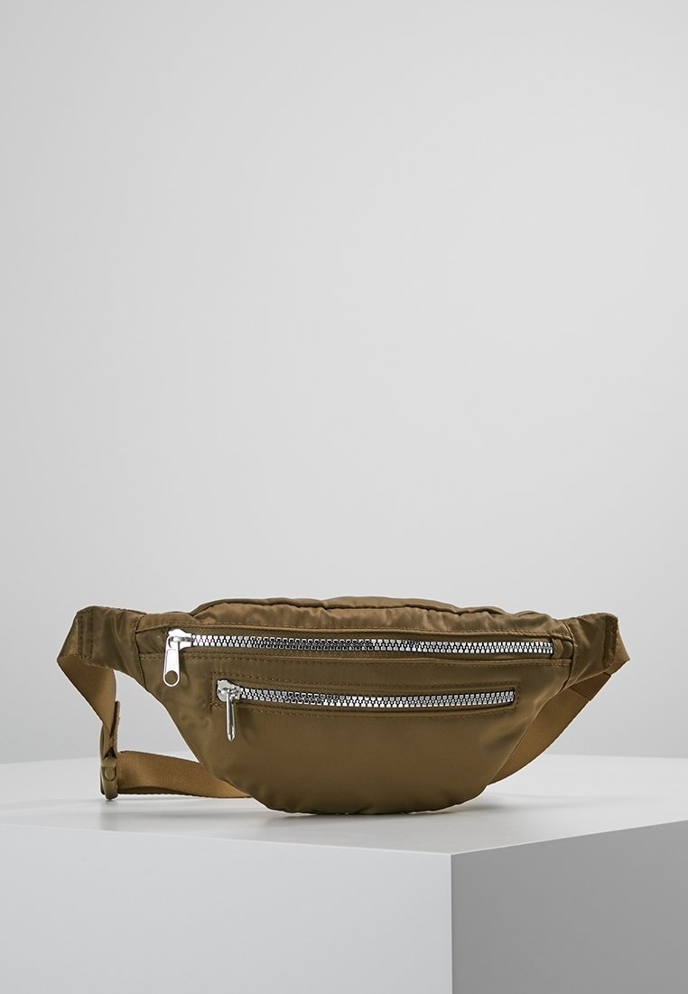 Sac banane marron avec deux poches zippées argentées et une sangle ajustable, posé sur une surface blanche devant un fond gris clair.