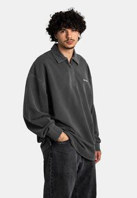 Langærmet polo i mørkegrå stof med ribbet tekstur, lynlås krave og broderet logo på brystet. Båret med sorte jeans.