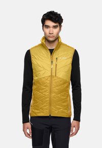 Gelbe gestepfte Weste mit Reißverschlusstaschen und Stehkragen, kombiniert mit einem schwarzen Langarmshirt. Strukturierter Stoff mit Zickzackmuster.