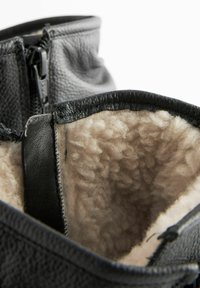 Musta nahkasaapas nilkkamittaisella, tekstuurilla varustetulla pinnalla, villavuorella ja vetoketjukiinnityksellä. Pehmeä, beige sisustus kontrastoi ulkopinnan kanssa.