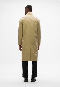 Khaki regenjas met een rechte snit, halflang, voorzien van een rugventilatie, gecombineerd met zwarte broeken en schoenen. Glad, waterafstotend materiaal.