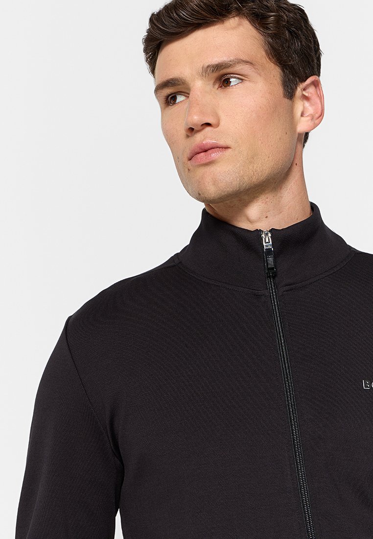 Giacca in fleece nera con zip e colletto alto, tessuto testurizzato e un piccolo logo sul lato sinistro del petto. Design semplice e funzionale.