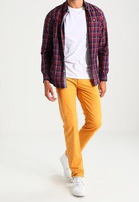 Rotes und marineblaues kariertes Hemd mit Knöpfen über einem weißen T-Shirt, kombiniert mit schmal geschnittenen gelben Hosen und weißen Sneakers.