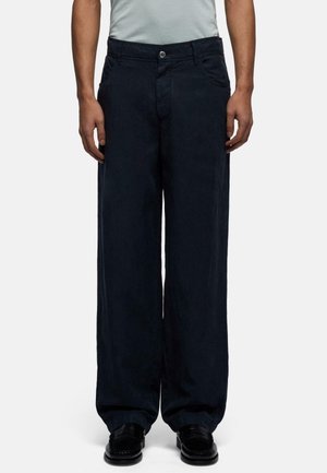Person trägt schwarze weite Hose mit vorderem Knopf und Taschen, kombiniert mit schwarzen Loafers und einem hellgrauen Hemd, das in die Hose gesteckt ist.