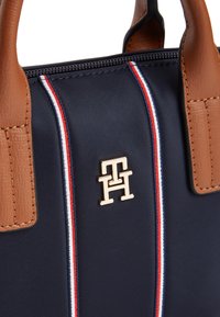 Ναυτικό νάιλον τσάντα με καφέ δερμάτινες λαβές, με χρυσό λογότυπο και κόκκινες, λευκές και μπλε κάθετες ρίγες ως διακοσμητικά στοιχεία. Κλείνει με φερμουάρ.