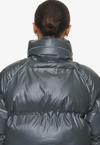 Grijze pufferjas met een glanzende afwerking, hoge kraag en geplooide secties. Kenmerkt zich door het "PEGADOR"-logo op de achterkant en contrasterende stikdetails.