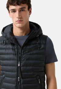 Schwarze Daunenweste mit Kapuze, ausgestattet mit Reißverschlusstaschen, einem gesteppten Textur und einem eleganten Design. Getragen über einem dunkelgrauen Shirt.