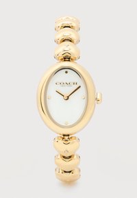 Χρυσό ρολόι χειρός Coach με οβάλ λευκό καντράν, χρυστά δείκτες, ελάχιστους δείκτες και βραχιόλι με συνδέσμους σε σχήμα καρδιάς με μικρά κρύσταλλα.