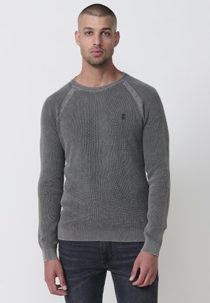 Suéter de punto gris con un patrón texturizado, cuello redondo y puños de canalé. Presenta un pequeño logotipo en el pecho. Combinado con jeans oscuros.