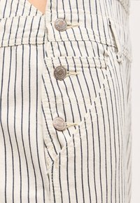 Κοντινή λήψη λευκών παντελονιών με λεπτές μαύρες κάθετες ρίγες, με τρία ασημένια μεταλλικά κουμπιά και μια μπροστινή τσέπη.