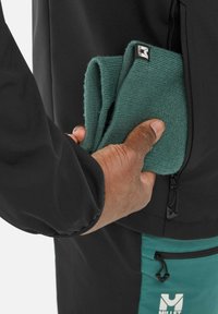 Veste noire avec une poche zippée, tenant un tissu vert plié. Le panneau latéral présente un accent teal et un logo de marque.