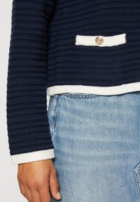 Haut à manches longues en tissu côtelé bleu marine avec bordure blanche et bouton doré, porté sur un jean en denim bleu clair sur le torse et le bras d'une personne.