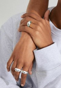 Anillos de metal plateado con una superficie lisa y pulida, presentando un diseño redondeado. Las manos están unidas, llevando un sudadera gris claro.