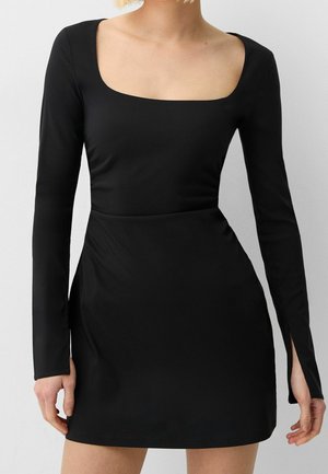 Frau trägt ein figurbetontes schwarzes Mini-Kleid mit langen Ärmeln, quadratischem Ausschnitt und dezenten Schlitzen an den Manschetten.