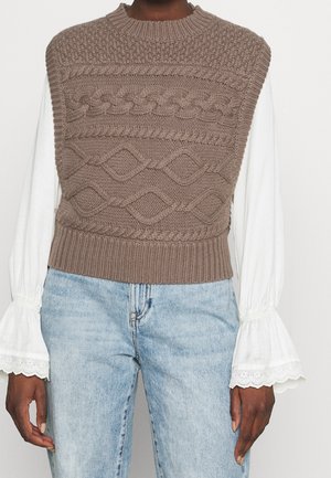 Pull sans manches marron tricoté avec des motifs torsadés, porté sur un chemisier blanc aux manches évasées en dentelle et un jean bleu clair.
