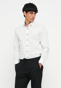 Witte blouse met lange mouwen, gemaakt van gladde stof. Voorzien van een klassieke kraag, blauwe knopen en een getailleerd ontwerp, gecombineerd met zwarte trousers.