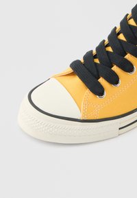 Κίτρινο καμβά sneaker με μαύρα κορδόνια, λευκό λαστιχένιο περίβλημα στα δάχτυλα και λευκή υφή σόλας πάνω σε ανοιχτό φόντο.