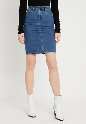 Kvinde iført en blå denim nederdel til knæet, sort langærmet top og hvide ankelstøvler, stående mod en ensartet lys baggrund.