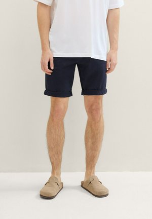 Granatowe bawełniane szorty z podwiniętymi mankietami, w połączeniu z jasnobrązowymi butami typu slip-on. Koszulka to luźna biała T-shirt.