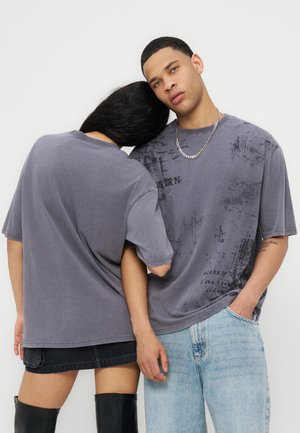 Zwei Personen tragen übergroße graue T-Shirts mit Motiv; eine lehnt den Kopf an die Schulter der anderen, die nach vorne schaut und die Hand in der Tasche hat.