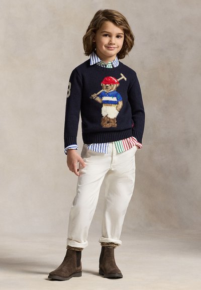 Niño pequeño de pie, vistiendo un suéter azul marino con un oso de peluche sosteniendo un mazo de polo, camisa de rayas, pantalones blancos y botas marrones sobre un fondo neutro.